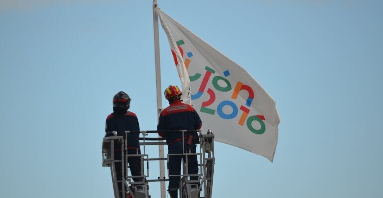 Dos miembros del equipo de bomberos colocan la bandera de Gijón 2016 en La Escalerona