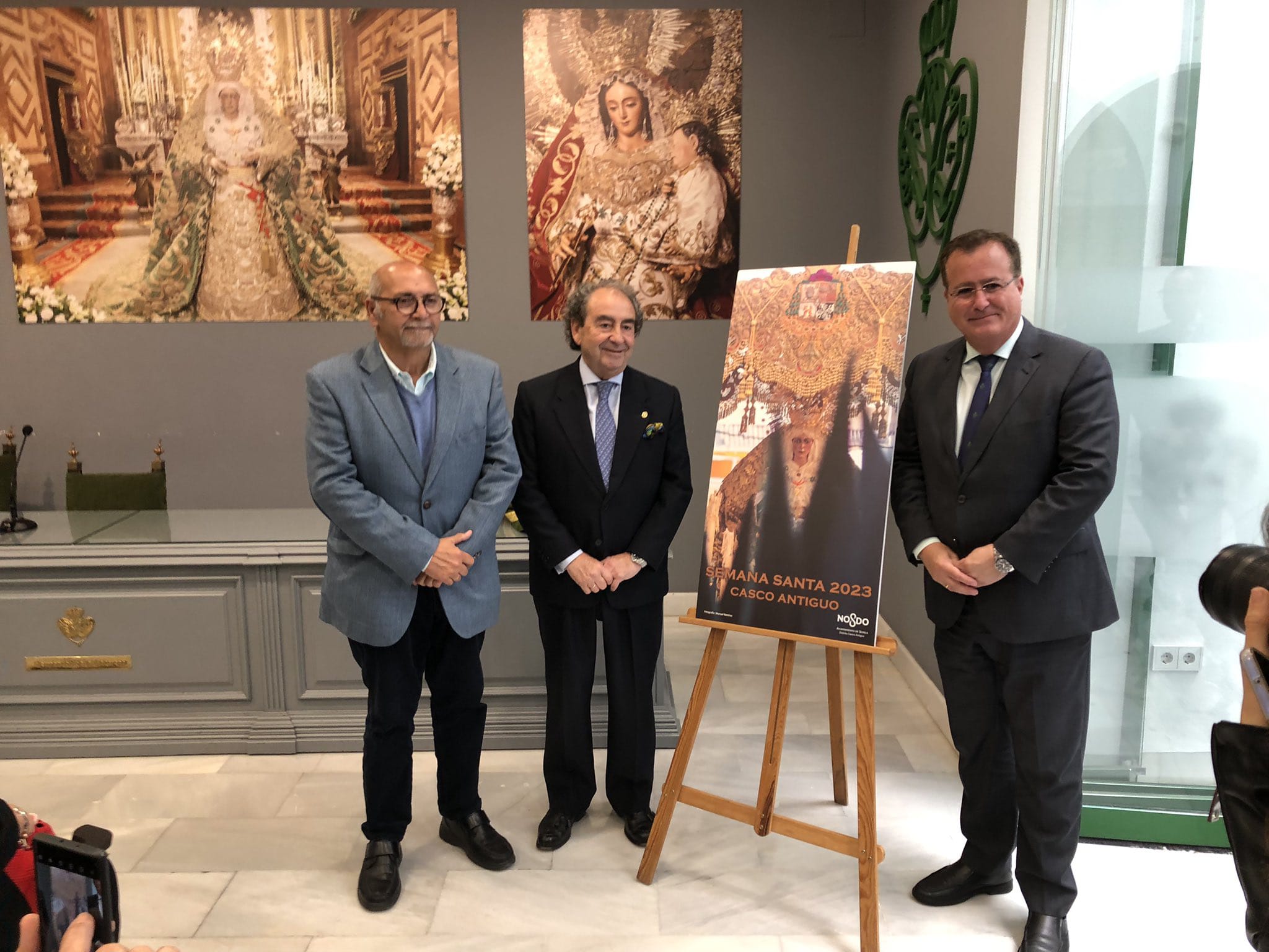 Junto al cartel del Distrito Casco Antiguo y de izquierda a derecha, el fotógrafo Manuel Ramírez Rubio, José Antonio Fernández Cabrero, hermano mayor de La Macarena; y el delegado municipal de Fiestas Mayores, Juan Carlos Cabrera