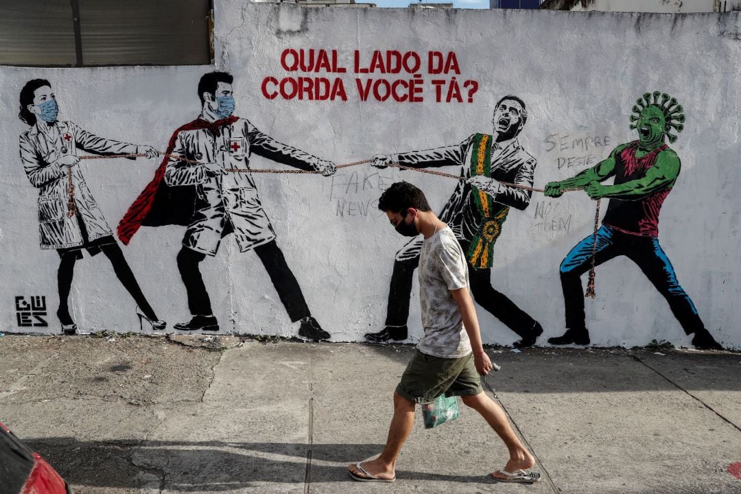 Un hombre pasa al frente de un graffiti que representa al presidente brasileño Jair Bolsonaro y una figura que representa el nuevo coronavirus COVID-19 tirando de una cuerda contra los trabajadores de la salud con un mensaje que dice: &quot;¿De qué lado de la cuerda estás?&quot;