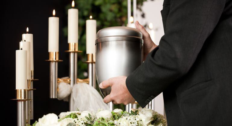 Imagen de un hombre sosteniendo una urna funeraria rodeado de velas y flores.