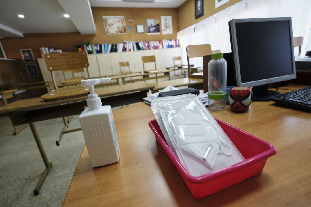 Mascarillas y gel desinfectante en la mesa del profesor de un aula donde se pueden observar los pupitres colocados y separados entre sí para el nuevo curso.