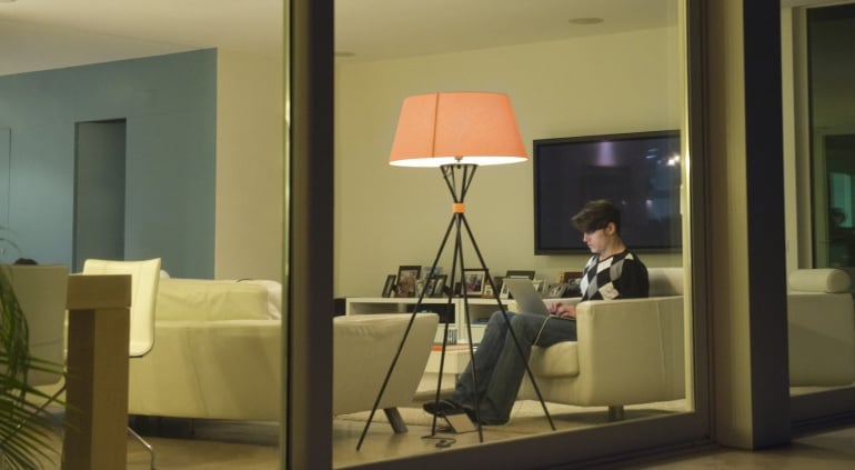 Vista a través de una ventana de un hombre utilizando un ordenador portátil con las luces del salón de su casa encendidas.