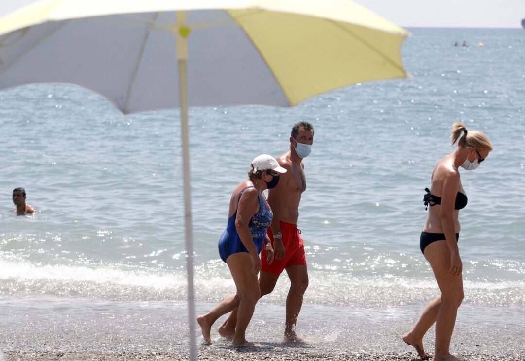 Bañistas con mascarilla pasean por la orilla de una playa.