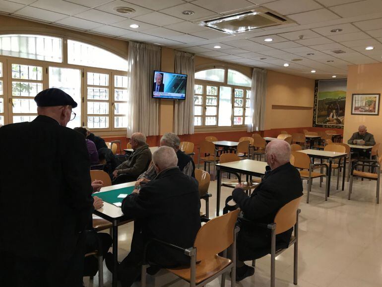 Varias personas en un club de jubilados jugando a las cartas y viendo la televisión.