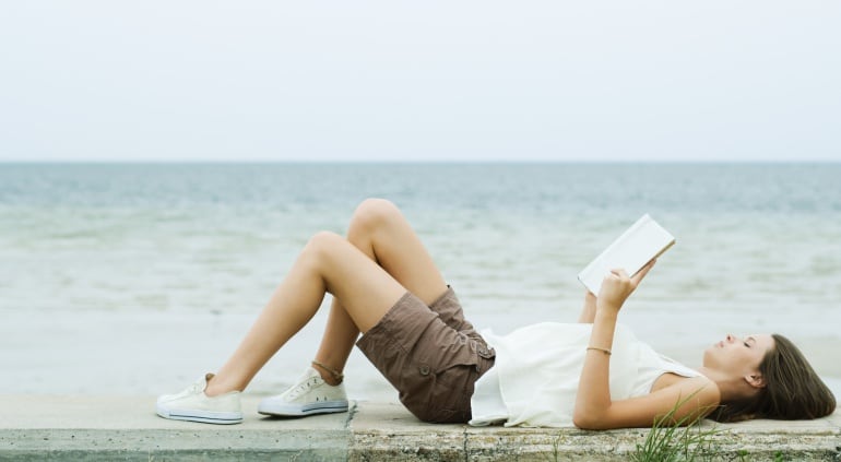 Una chica lee un libro tumbada en un muro frente al mar.