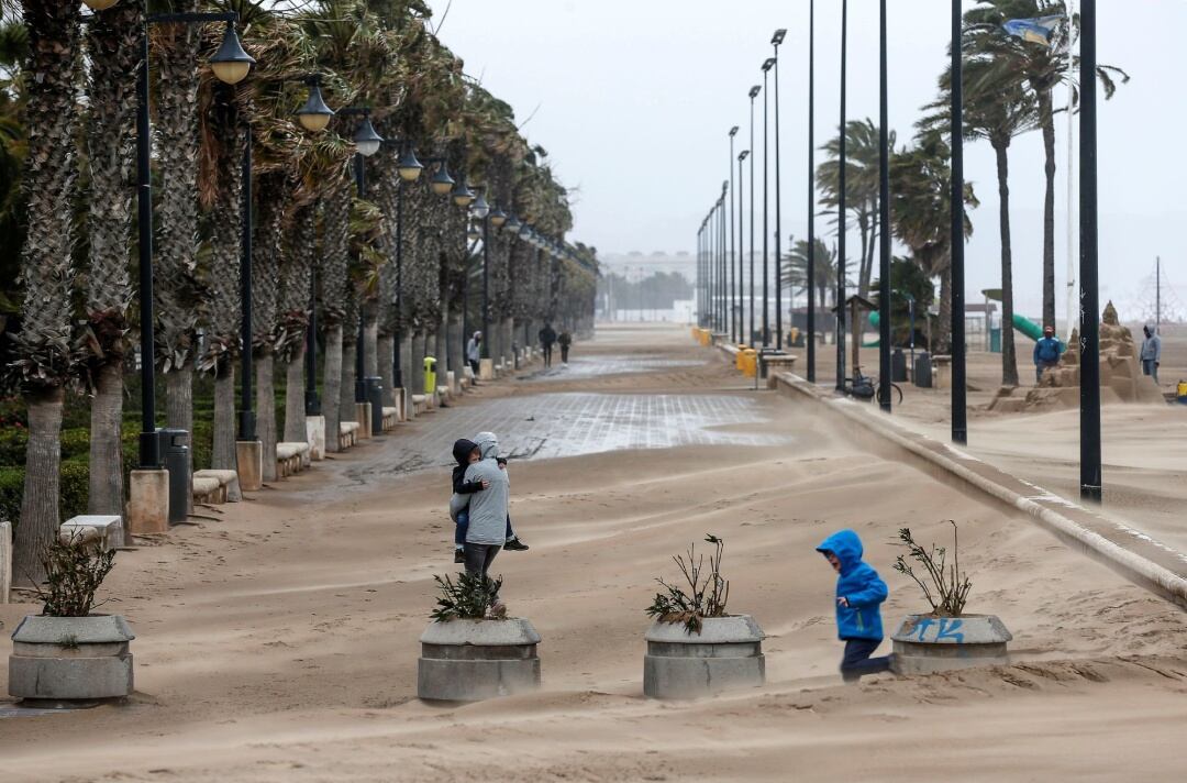 Una mujer cruza el paseo marítimo de Valencia cubierto en parte por la arena que arrastra el fuerte viento