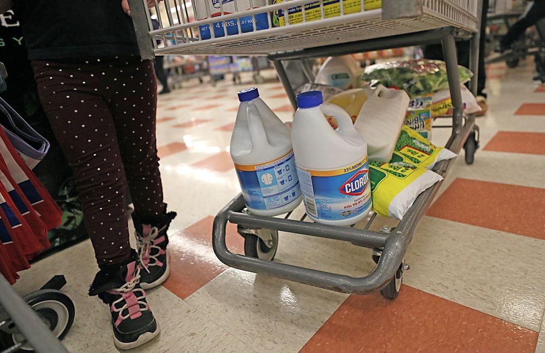 Una niña junto a un carro de compra en el que hay varias botellas de compuestos químicos con cloro.