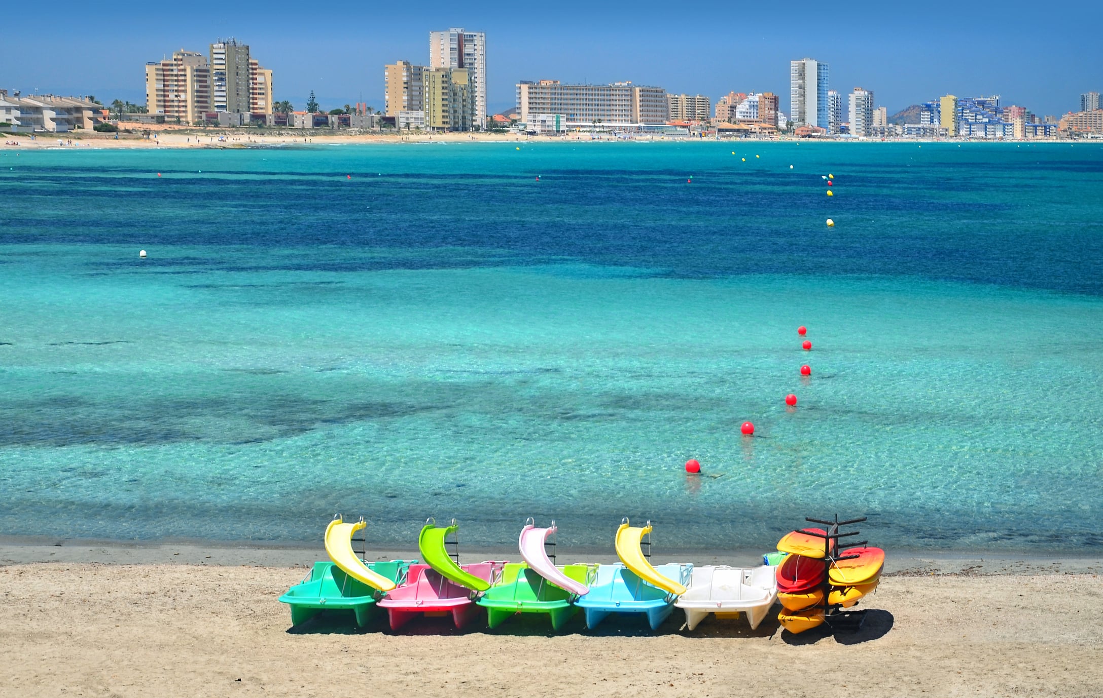 Un grupo de embarcaciones de colores amarradas en la arena con el fondo de las aguas turquesas de la playa de Levante, en verano, en Cabo de Palos, cerca de La Manga del Mar Menor, Murcia. Uno de los lugares más turísticos del levante español. Al fondo, el contraste de los grandes edificios de La Manga