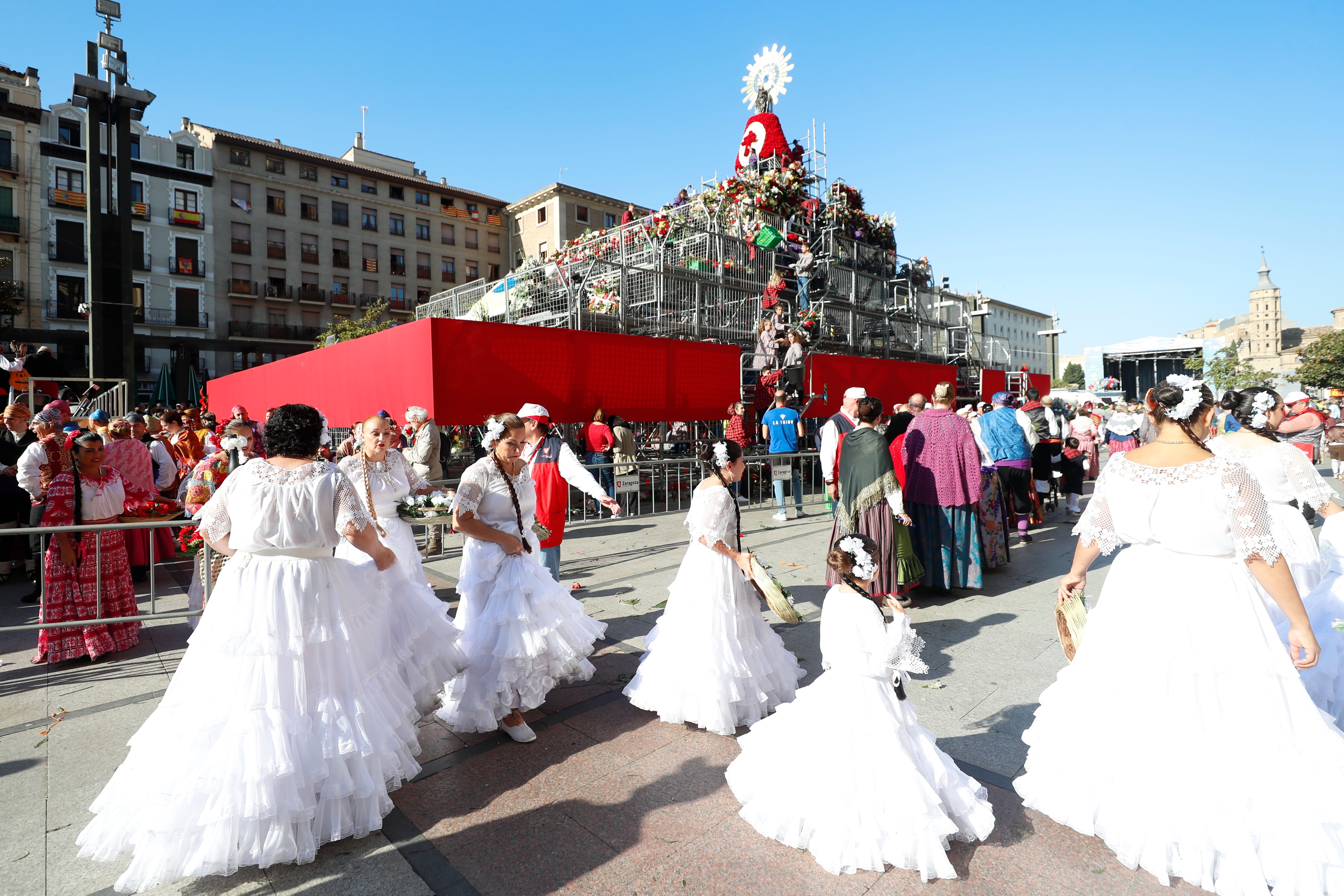 ZARAGOZA, 12/10/2022.- Varias personas bailan junto a la imagen de la Virgen del Pilar que luce ya las primeras flores en una estructura levantada en el centro de la plaza de Zaragoza que lleva su nombre, una ofrenda llena de ilusión que se espera de récord tras dos años de pandemia, con más grupos inscritos que en 2019 y la previsión de alcanzar alrededor de siete millones de flores. EFE/ Javier Cebollada
