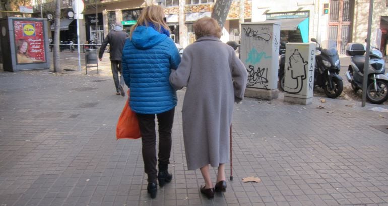 Una anciana camina ayudada por una asistente.