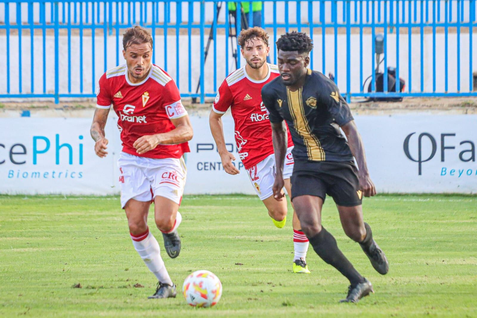 Angong, jugador del Intercity, controla el balón ante dos futbolistas del Murcia