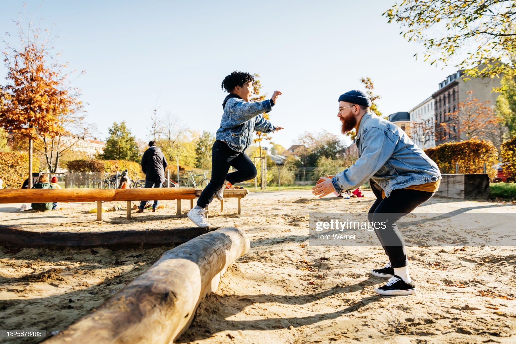 A young boy leaping into his fathers arms from a log while messing around in a playground at the park together.