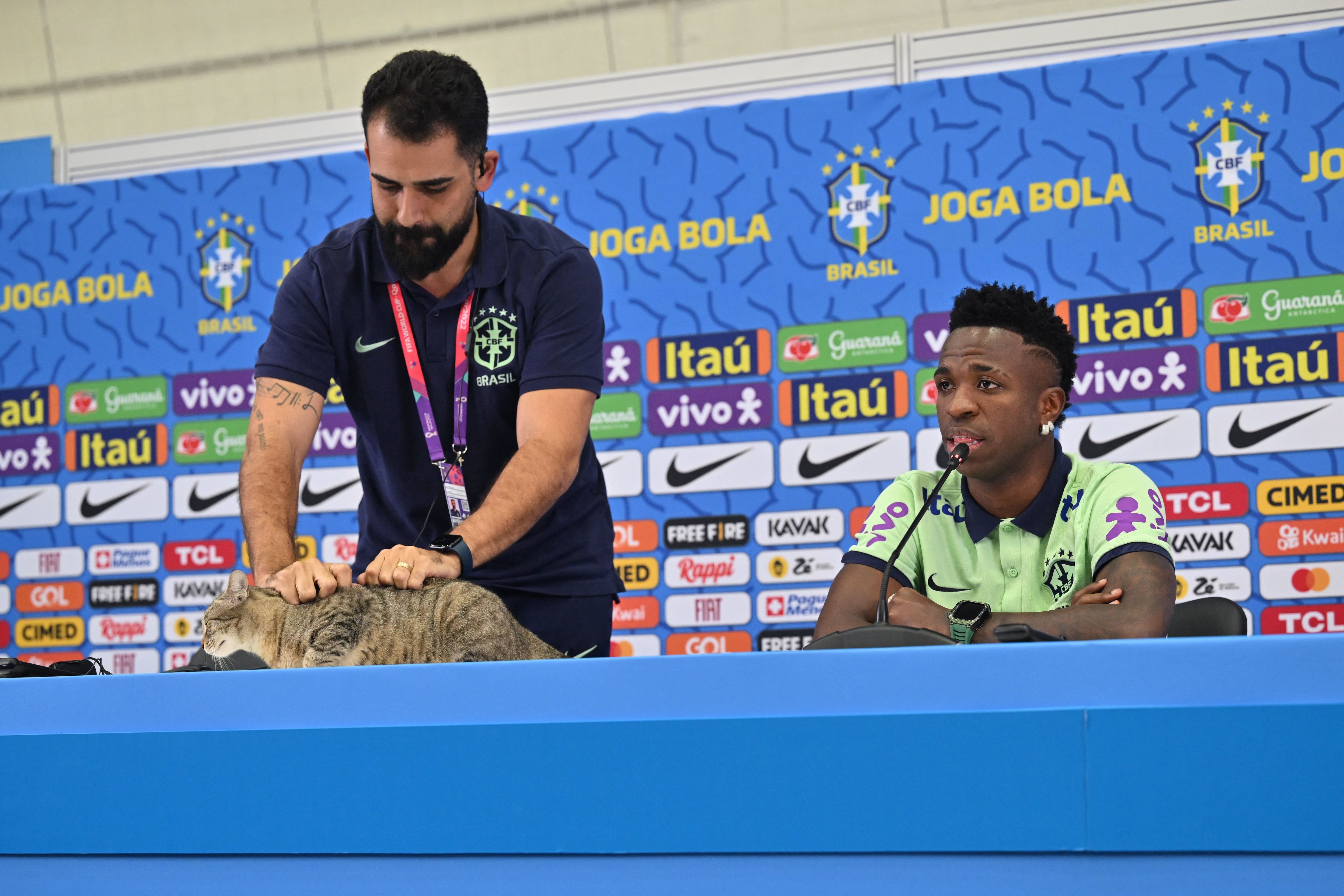 Un trabajador de prensa de Brasil agarra un gato para bajarlo de la mesa mientras Vinícius continúa en rueda de prensa