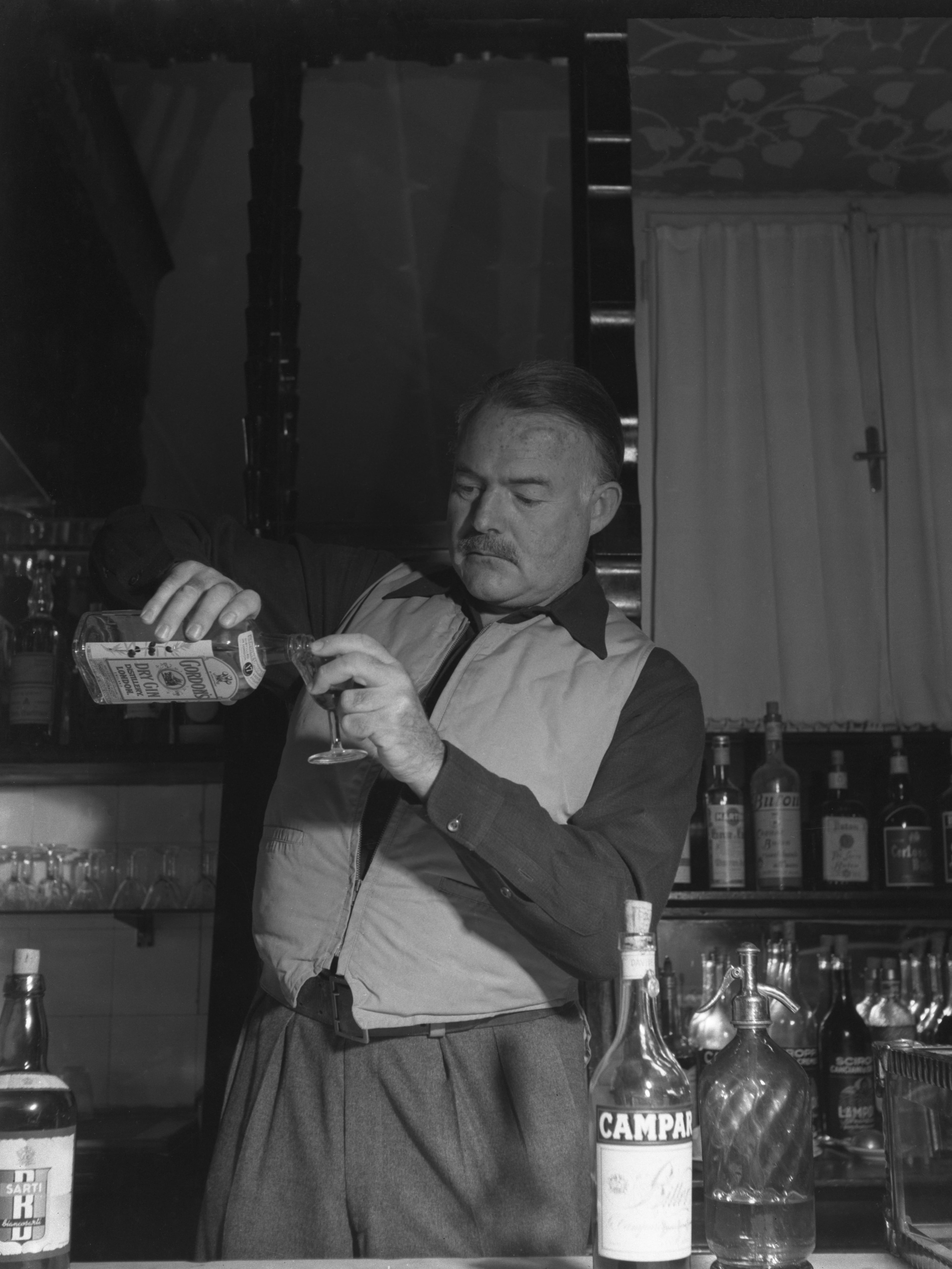 American writer Ernest Hemingway wearing a hunter waistcoat, standing behind a bar counter and pouring gin from a bottle of Gordon&#039;s, other alcoholic bottles around him, Cortina d&#039;Ampezzo, 1948