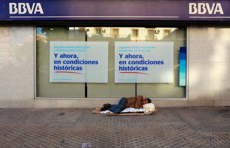 Un hombre duerme sobre cartones delante de una sucursal bancaria en Sevilla