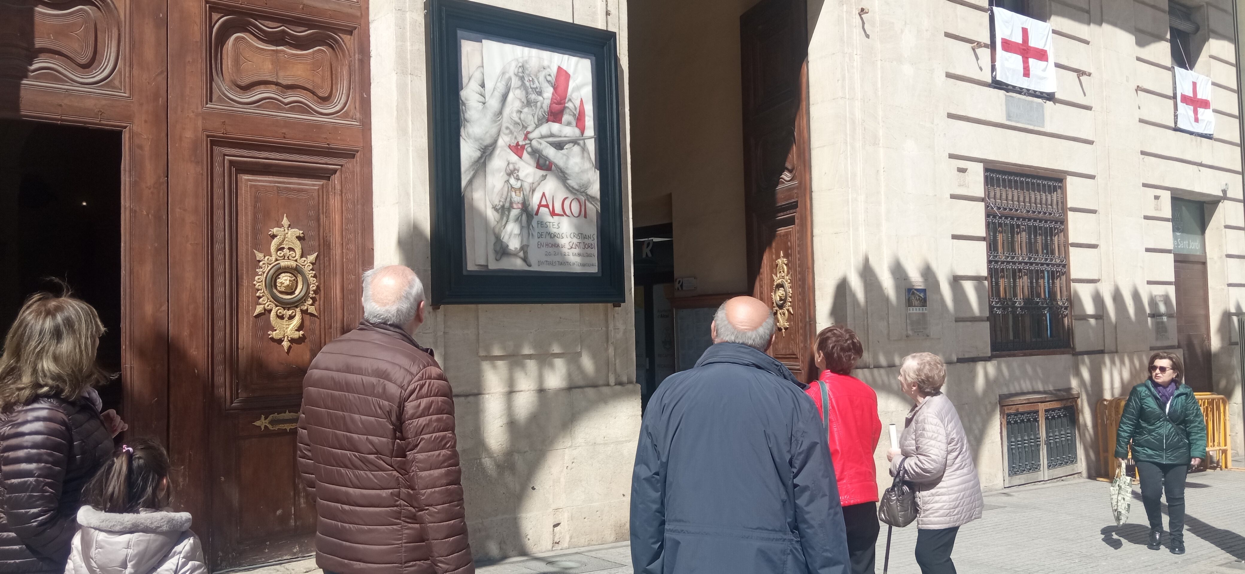 Ciudadanos viendo el cartel de las Fiestas de 2024, obra de Joan Castejón, que luce en la fachada del Ayuntamiento de Alcoy.