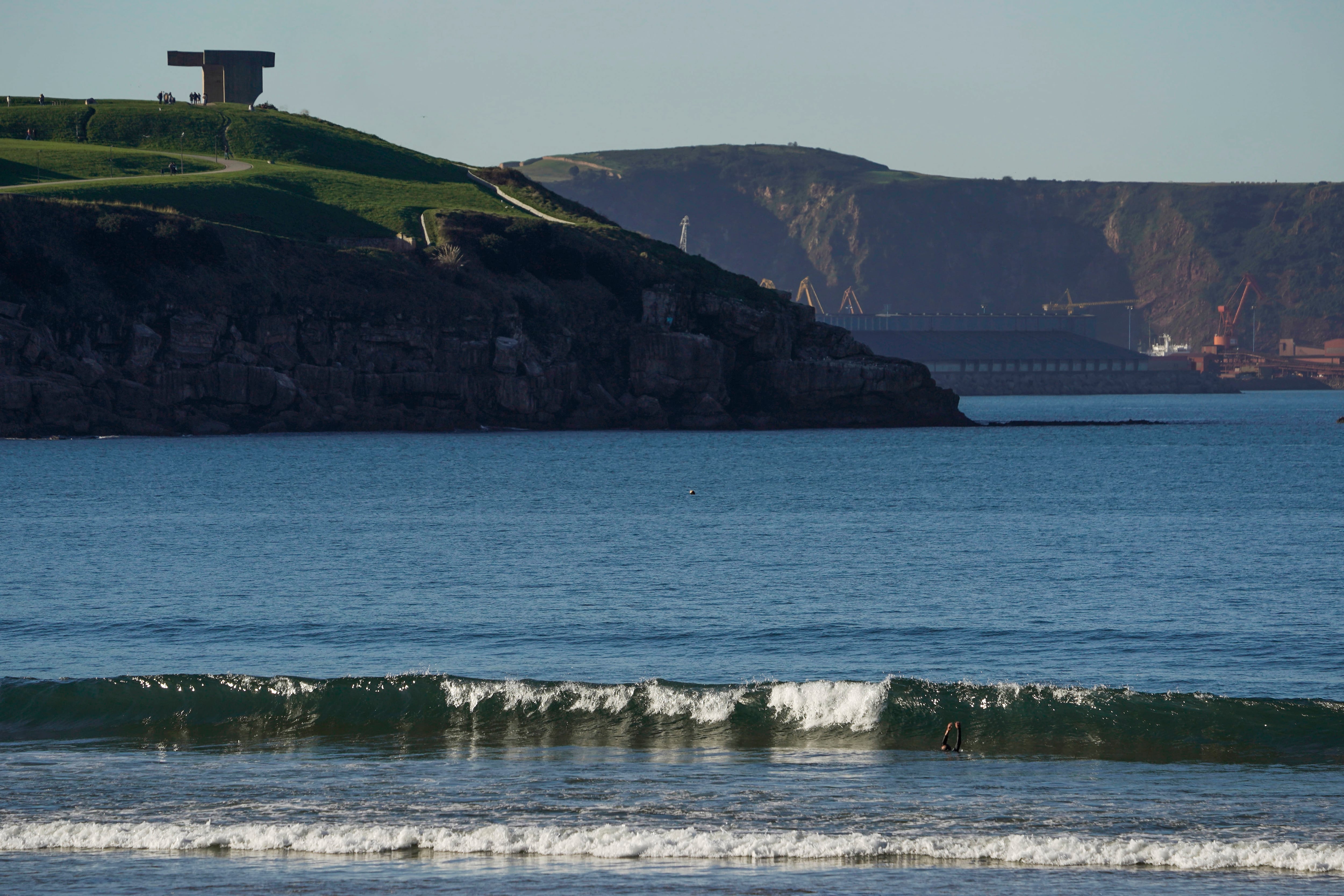 GIJON (ASTURIAS), 18/01/2025.- Un bañista entra en una ola en la playa de San Lorenzo con el Elogio del Horizonte al fondo este sábado, en Gijón. La Agencia Estatal de Meteorología (Aemet) prevé para mañana, sábado, en Asturias cielo poco nuboso y temperaturas estables. Heladas débiles en el interior, algo más fuertes en la cordillera Cantábrica. Viento flojo del sur. EFE/Paco Paredes
