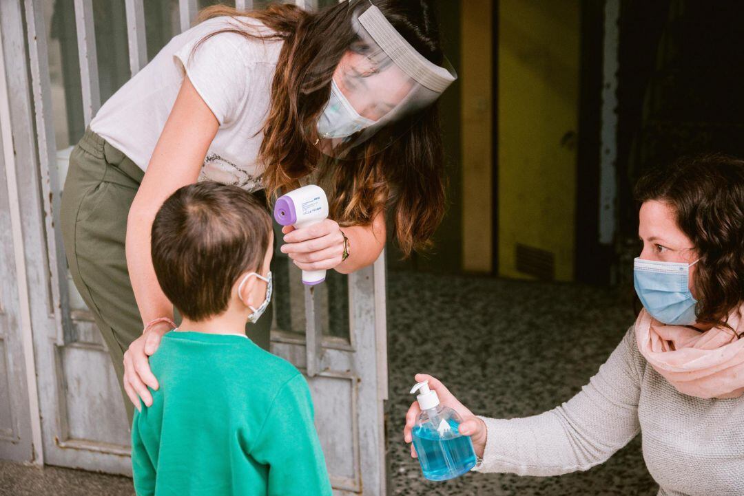 Dos trabajadoras de un centro educativo toman la temperatura y aplican gel hidroalcohólico en las manos a un niño.