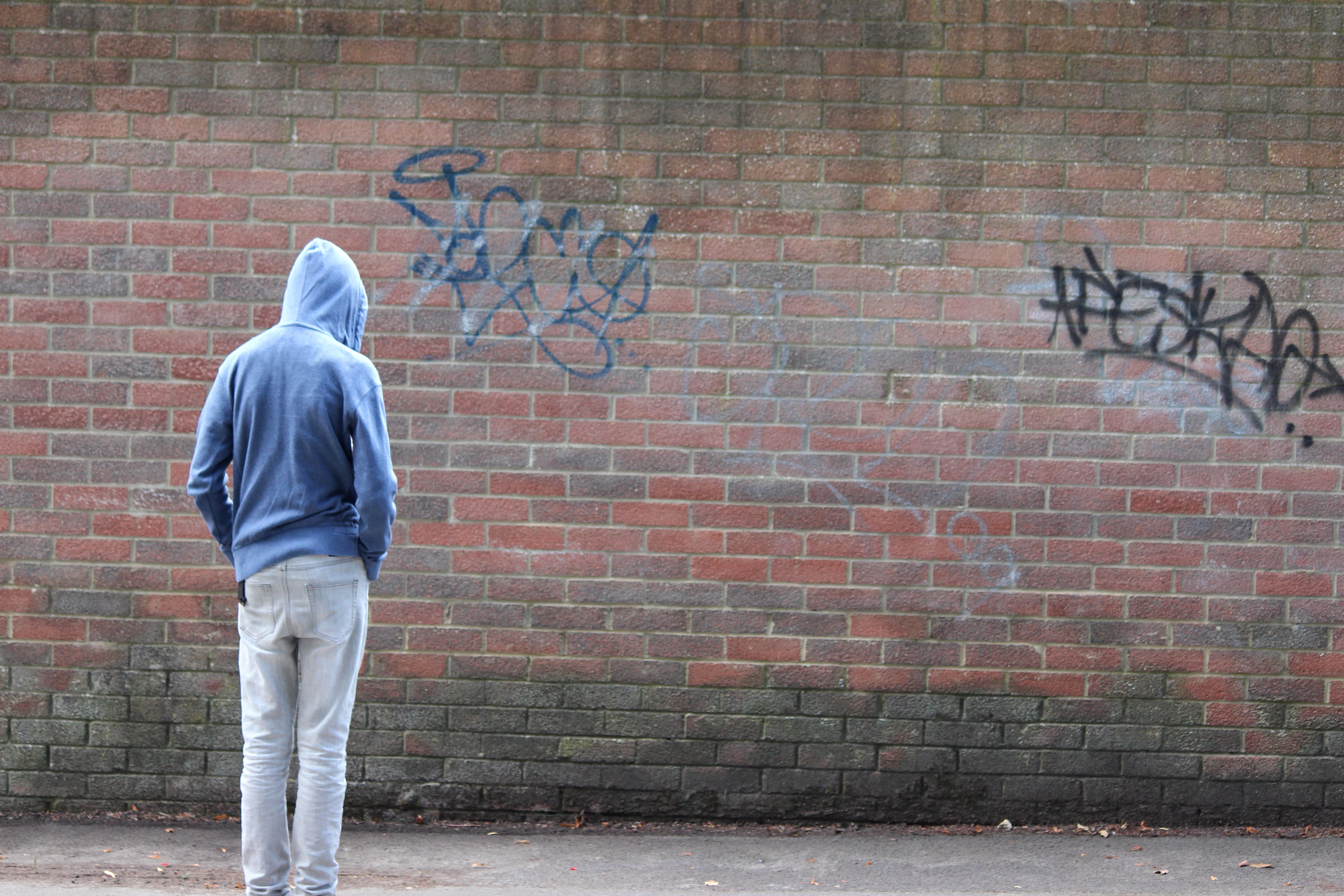 Photo showing a teenage boy / youth wearing a blue hoodie and standing beside a brick wall, in a run-down part of the city.