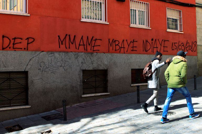 Dos personas pasan delante de una pintada en el madrileño barrio de Lavapiés, en memoria del &quot;mantero&quot; senegalés de 35 años que el jueves murió tras sufrir una parada cardiorrespiratoria