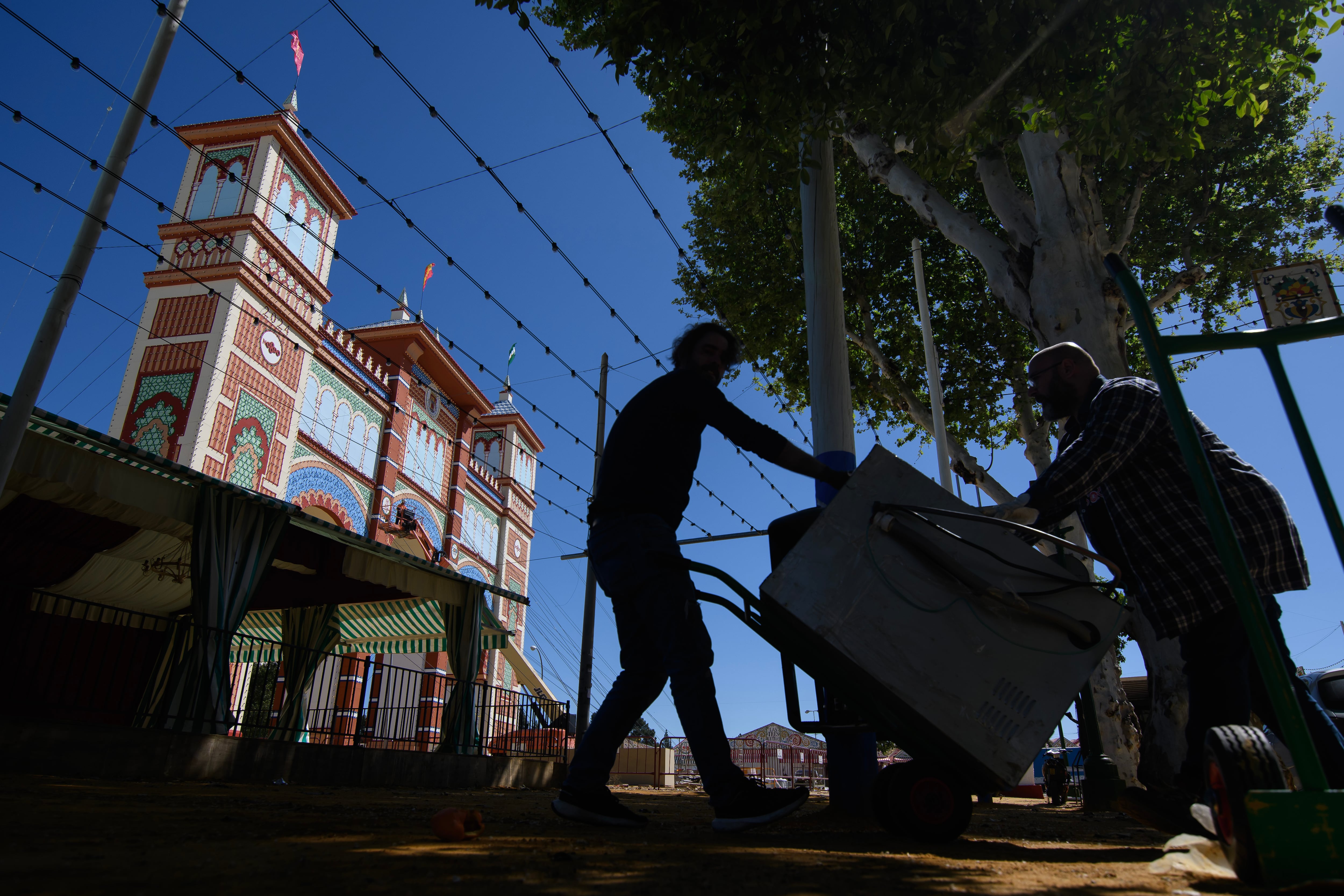 SEVILLA, 09/04/2024.- Dos hombres transportan este martes una nevera para una de las casetas del recinto de la Feria de Abril de Sevilla, que arranca el próximo fin de semana. EFE/ Raúl Caro
