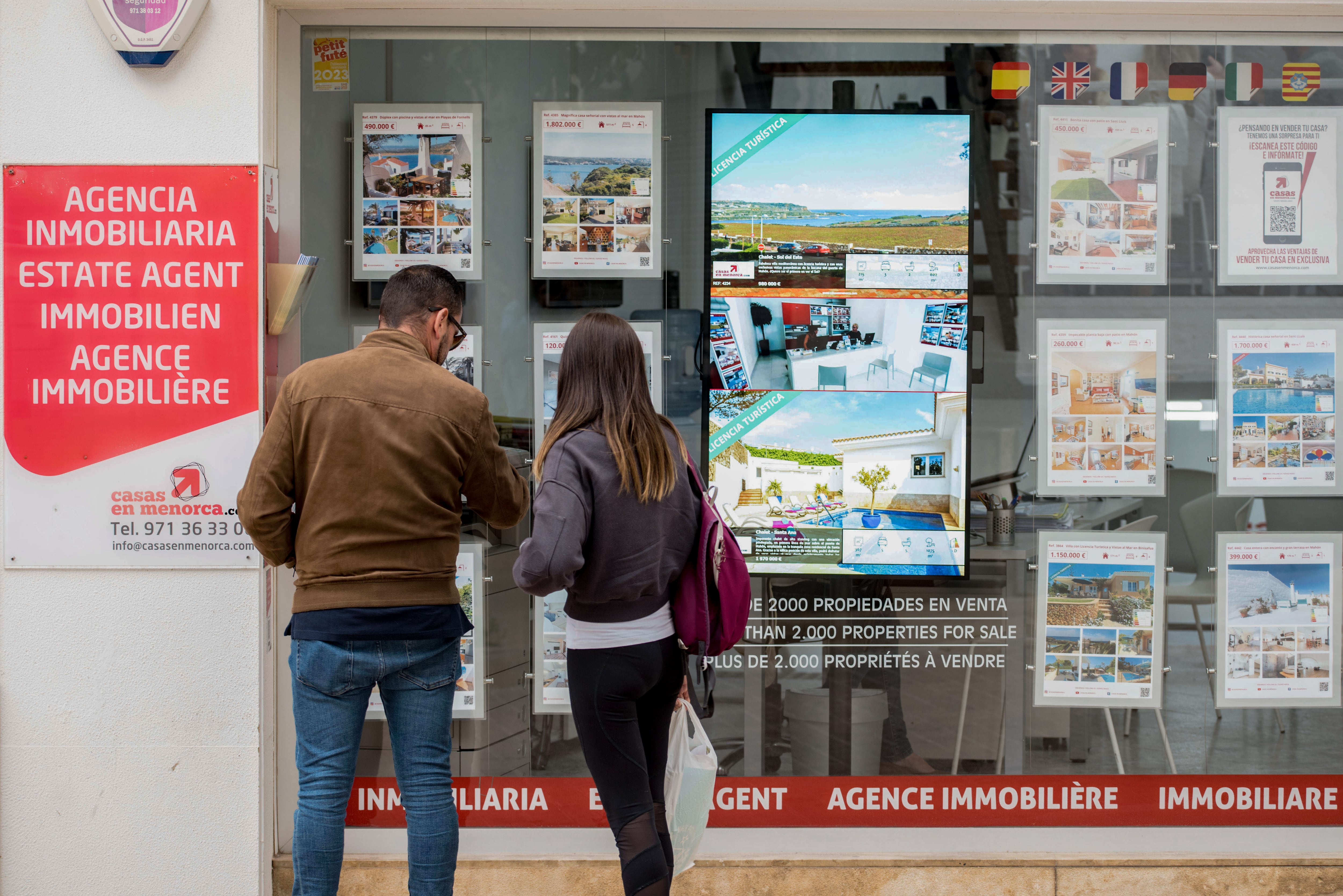 Una pareja se detiene a observar las propiedades en venta anunciadas en el escaparate de una agencia inmobiliaria de Menorca.