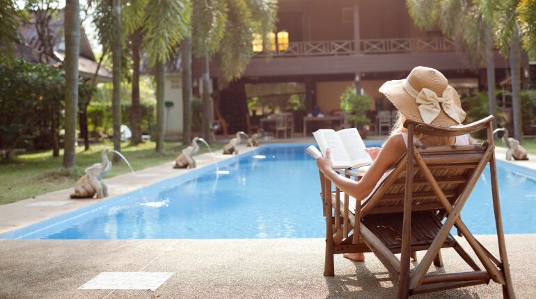 Imagen de una mujer leyendo en una tumbona frente a la piscina de una mansión. 