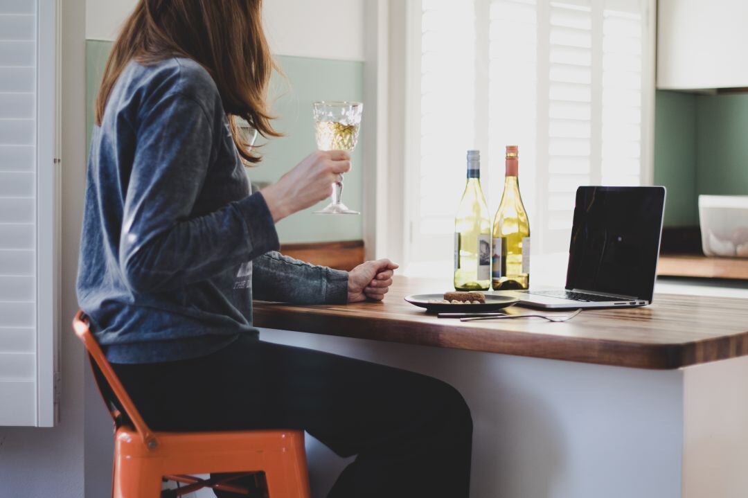 Una mujer sujeta una copa de vino junto a dos botellas de la misma bebida en un domicilio, frente a un ordenador. 