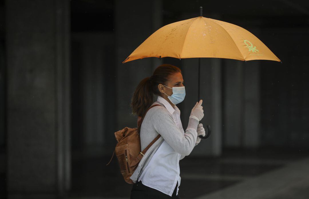 Una mujer protegida con mascarilla y guantes se resguarda de la lluvia bajo su paraguas.