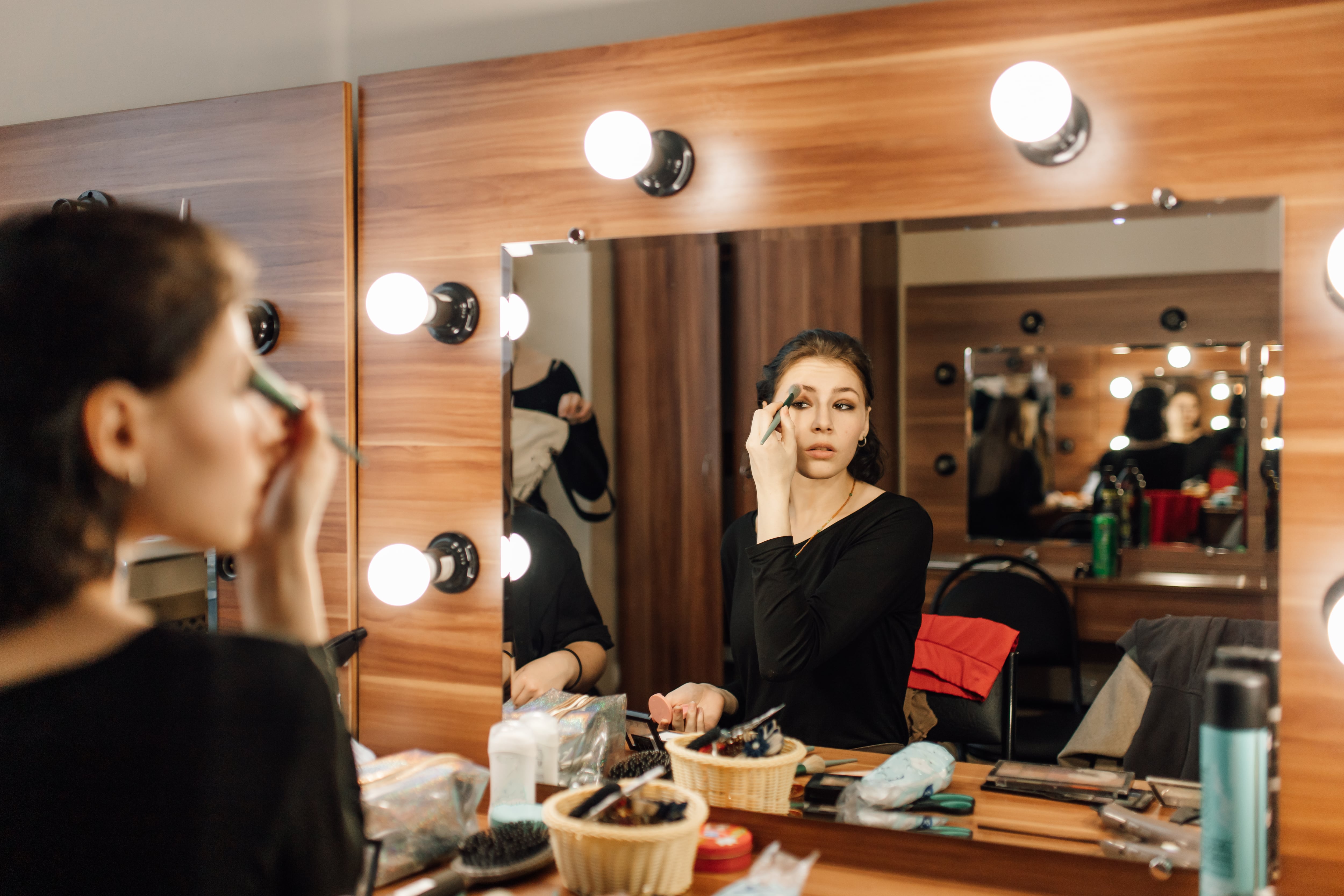 Confident young female performer with brown hair in long-sleeve black costume applying powder looking at reflection in mirror while sitting at dressing table getting ready for student drama club premiere event