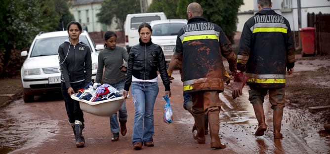 Tres mujeres jóvenes que cargan sus pertenencias encuentran a un par de bomberos a su paso por una calle cubierta de lodo rojo, en Devecser, a unos 164 kilómetros de la capital, Budapest. La ruptura de un dique en la localidad de Ajka produjo el vertido tóxico de un material conocido como &quot;barro rojo&quot;, una sustancia química muy tóxica, corrosiva y alcalina, lo que ha resultado en la muerte de cuatro personas