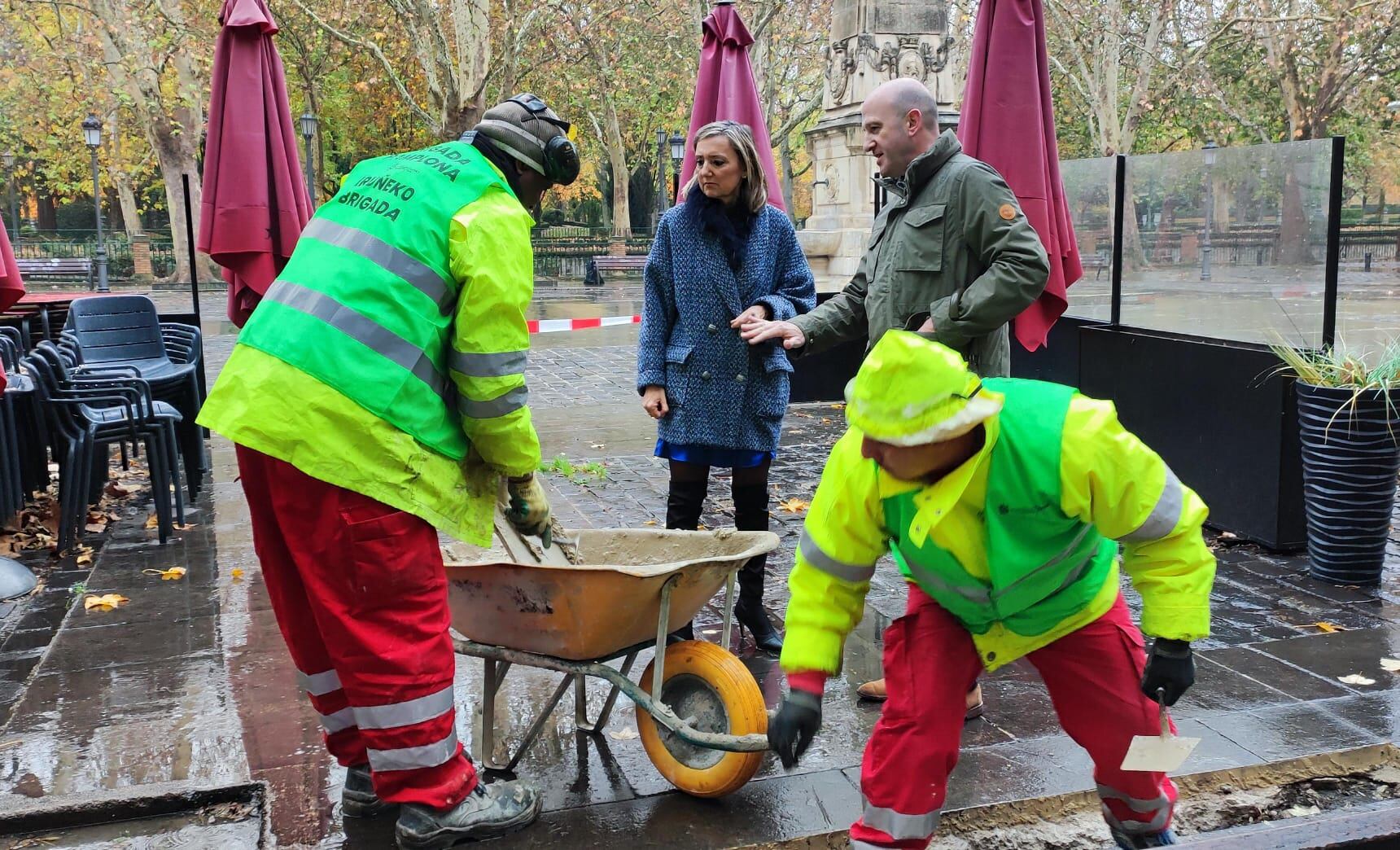 Dos operarios de la 'Brigada Pamplona' trabajan en unos desperfectos del pavimento en la plaza de Recoletas de Pamplona ante la mirada de la alcaldesa de la ciudad, Cristina Ibarrola (UPN) y el director del área de Conservación Urbana, Óscar Esquíroz.