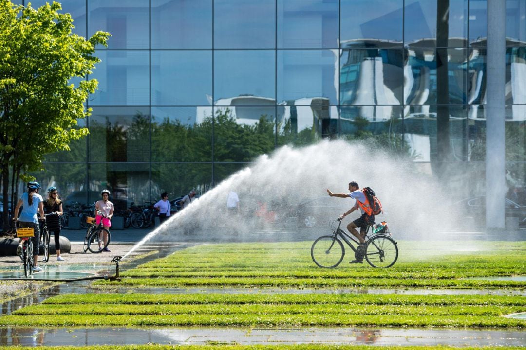 Un hombre en bicicleta se refresca con el agua de unos aspersores.