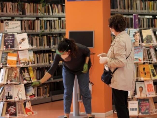 Una bibliotecaria recoge con guantes el libro elegido por una mujer para llevárselo a casa en préstamo. Ambas van protegidas con mascarilla y mantienen la distancia recomendada.