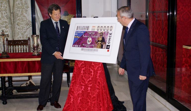 El hermano mayor de La Paz, Santiago Arenado, a la izquierda de la imagen, muestra el cupón del 75 aniversario de la bendición del Señor de la Victoria, junto al delegado de la ONCE en Andalucía, José Antonio Toledo