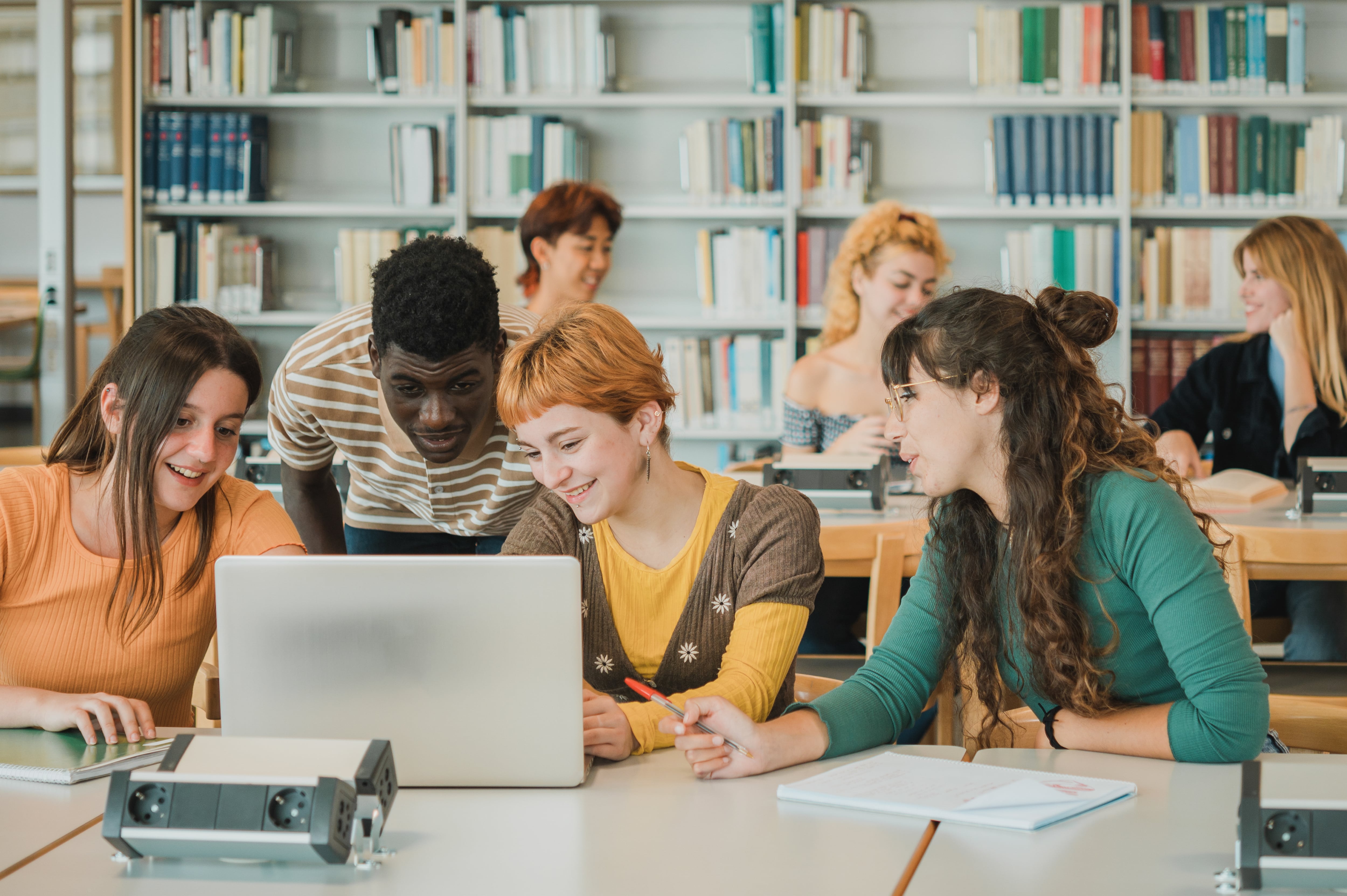 Group of multiethnic classmates in casual clothes smiling and analyzing data on netbook while gathering near table and working on project in university library