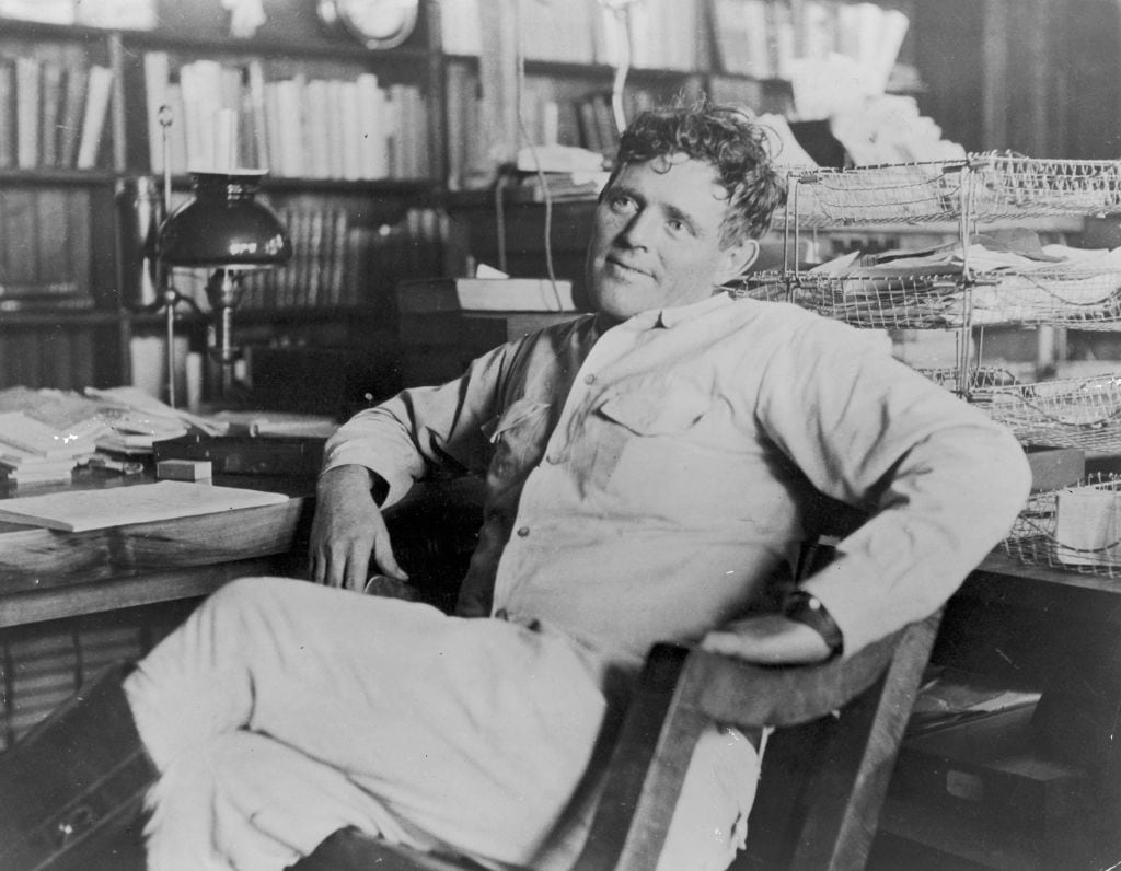 American author Jack London (1876-1916) reclines next to his desk in a wooden chair, smiling, his legs crossed.   (Photo by Hulton Archive/Getty Images)