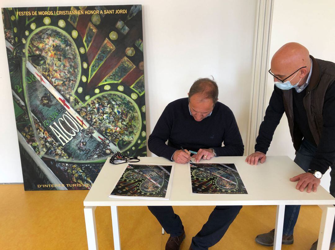 El pintor Luis Sanus firmando una de las postales de su cartel ante el director del Economato, Juanjo Garrido, con el cartel al fondo