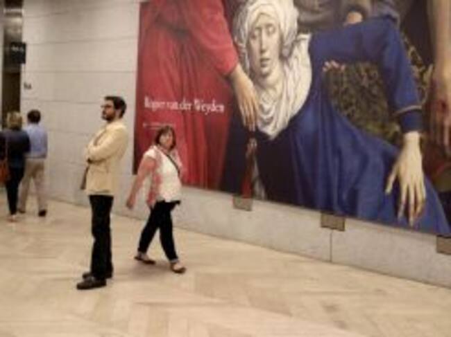 Dos espectadores junto a un cartel con un fragmento del "Descendimiento de la cruz" del flamenco Rogier van der Weyden, en el museo de El Prado.