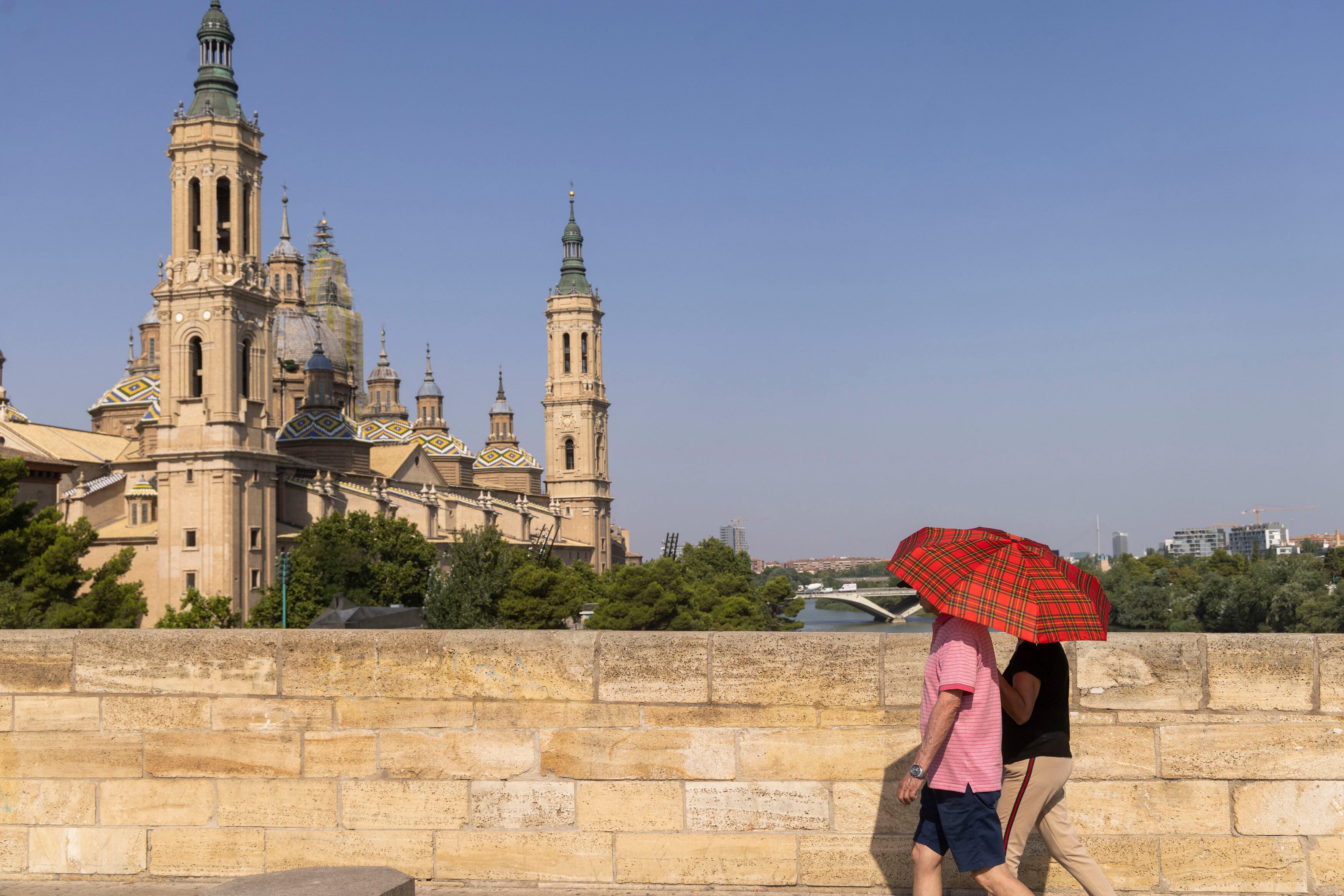 ZARAGOZA, 07/08/2025.- Una pareja cubierta con un paraguas para protegerse del sol pasa este jueves, por el puente de Piedra en Zaragoza. La ribera del Ebro y la Ibérica zaragozana se encuentran este jueves en alerta naranja por calor. EFE/ Toni Galán
