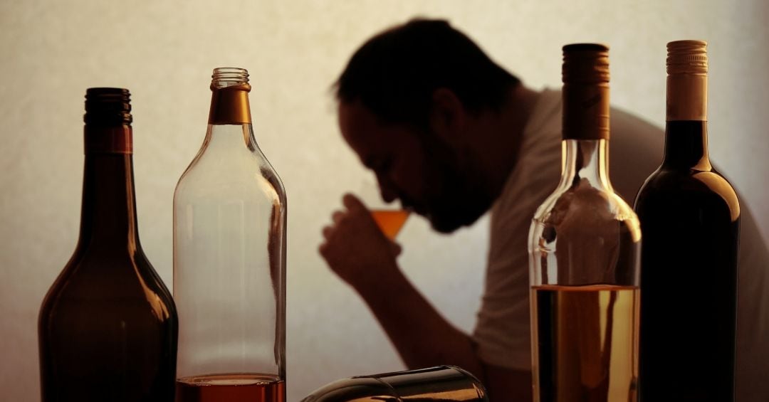 Un hombre bebe de un vaso entre botellas de alcohol.