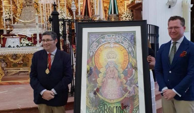 El presidente de la Hermandad Matriz de Almonte, Juan Ignacio Reales, a la izquierda, y el pintor nazareno Juan Miguel Martín Mena flanquean en el Santuario de la Blanca Paloma el cartel del centenario de la coronación canónica de la Virgen del Rocío