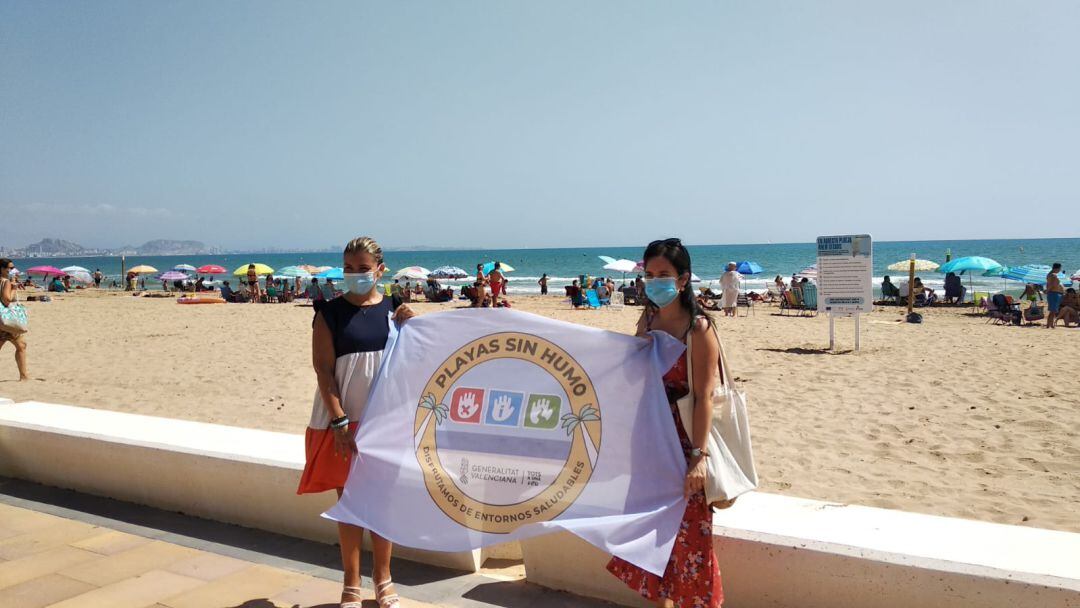 Mari Carmen Sánchez (d) y una técnico de la concejalía, con la bandera de playa sin humo de Urbanova