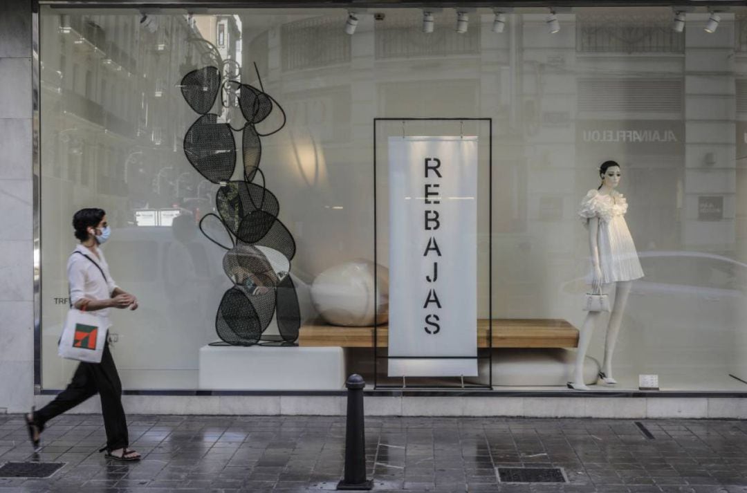 Una mujer protegida con mascarilla pasa al lado del escaparate de una tienda donde se observan carteles indicativos de rebajas en València