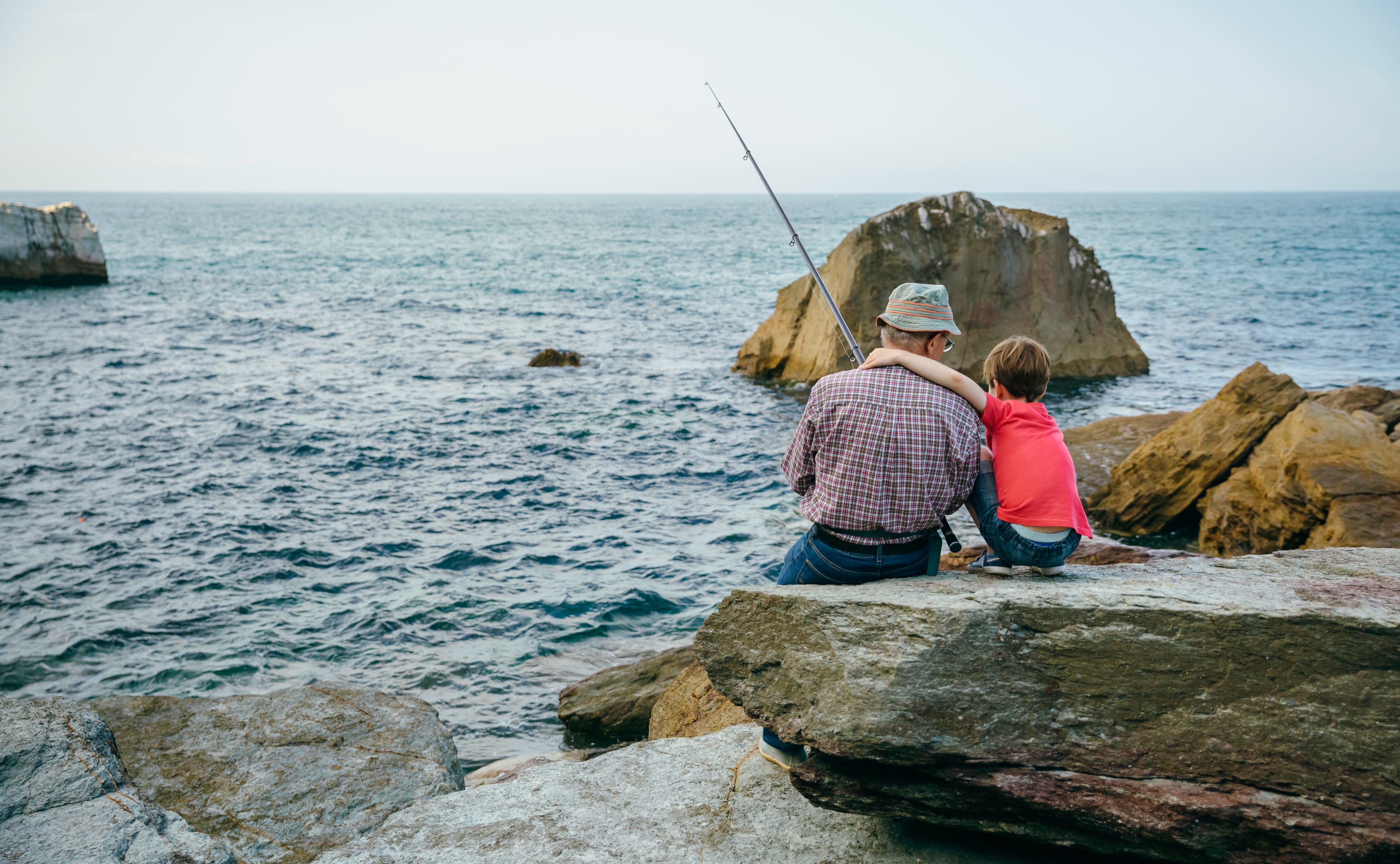 Spain, Asturias, Back view of unrecognizable child crouched down hugging to senior man while fishing with a rod into the sea sitting over big rock