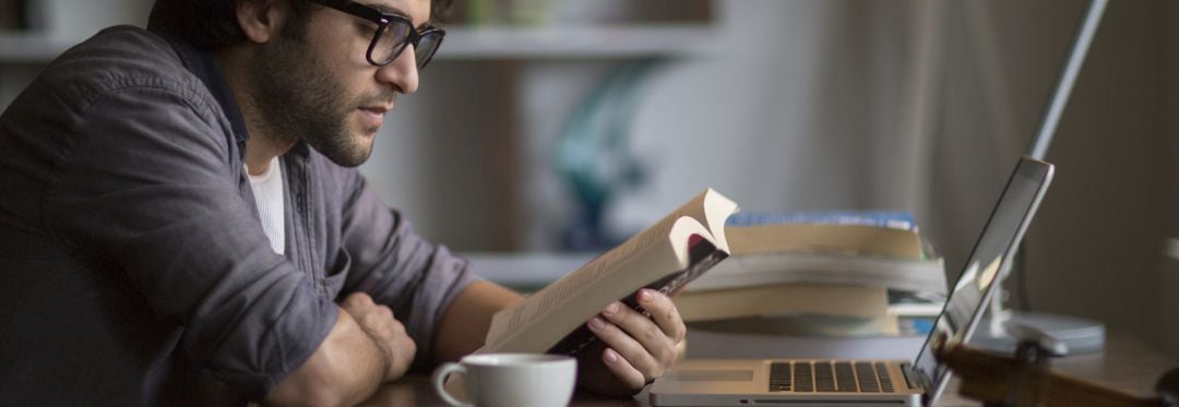 Hombre leyendo un libro frente a su ordenador