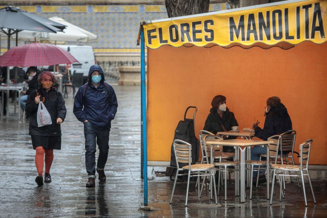 Dos personas en la terraza de un bar protegidas de la lluvia por un toldo de un puesto de flores en València.