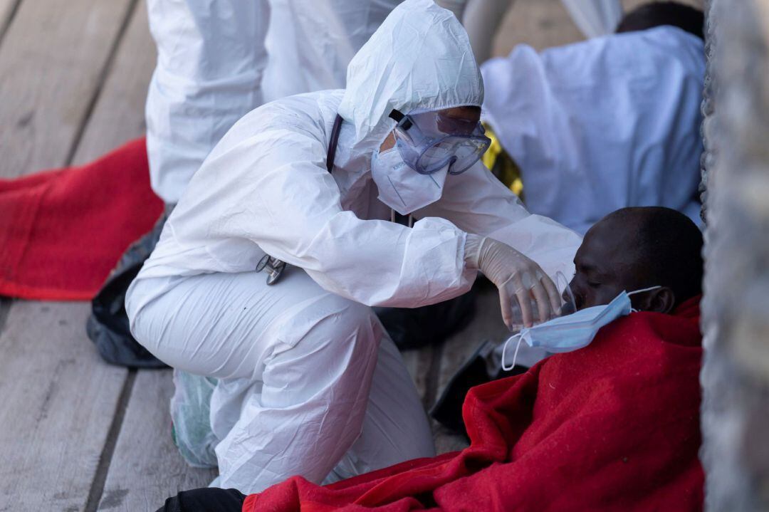 Miembros de los servicios sanitarios y de emergencia atienden, protegidos por trajes de seguridad del protocolo de prevención de la covid-19, a uno de los inmigrantes llegados a la costa