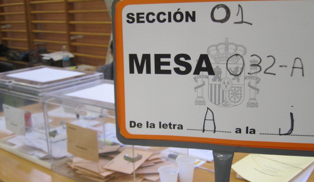 Cartel de una mesa electoral con las urnas al fondo.