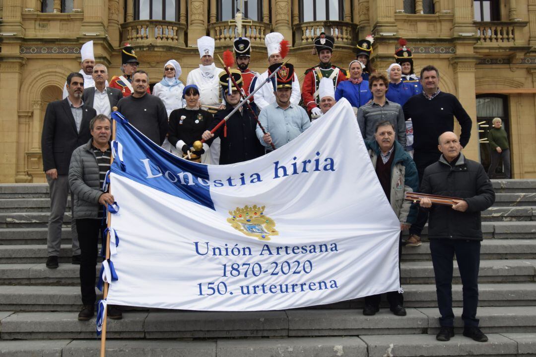 El tambor mayor de la Unión Artesana y de Gaztelubide, centro, junto a representantes políticos y otros protagonistas del día de San Sebastián, posan con la bandera que ondeara en la plaza de la Constitución.